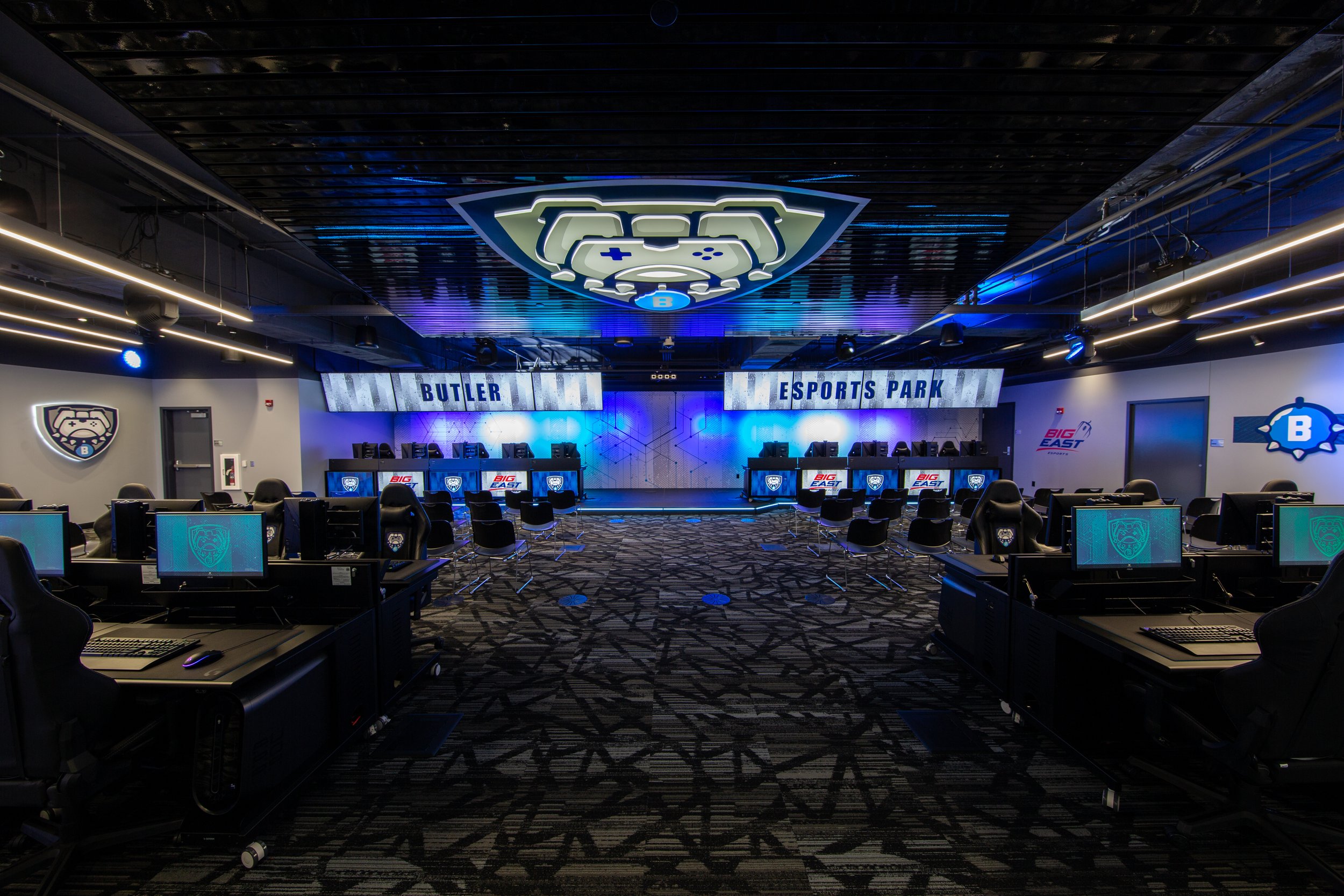 Interior of an esports gaming arena featuring rows of computer stations, gaming chairs, and illuminated signage with the text 'Butler Esports Park'. The ceiling has a large, illuminated logo resembling a game controller. Walls display additional logos and lighting creates a vibrant atmosphere.