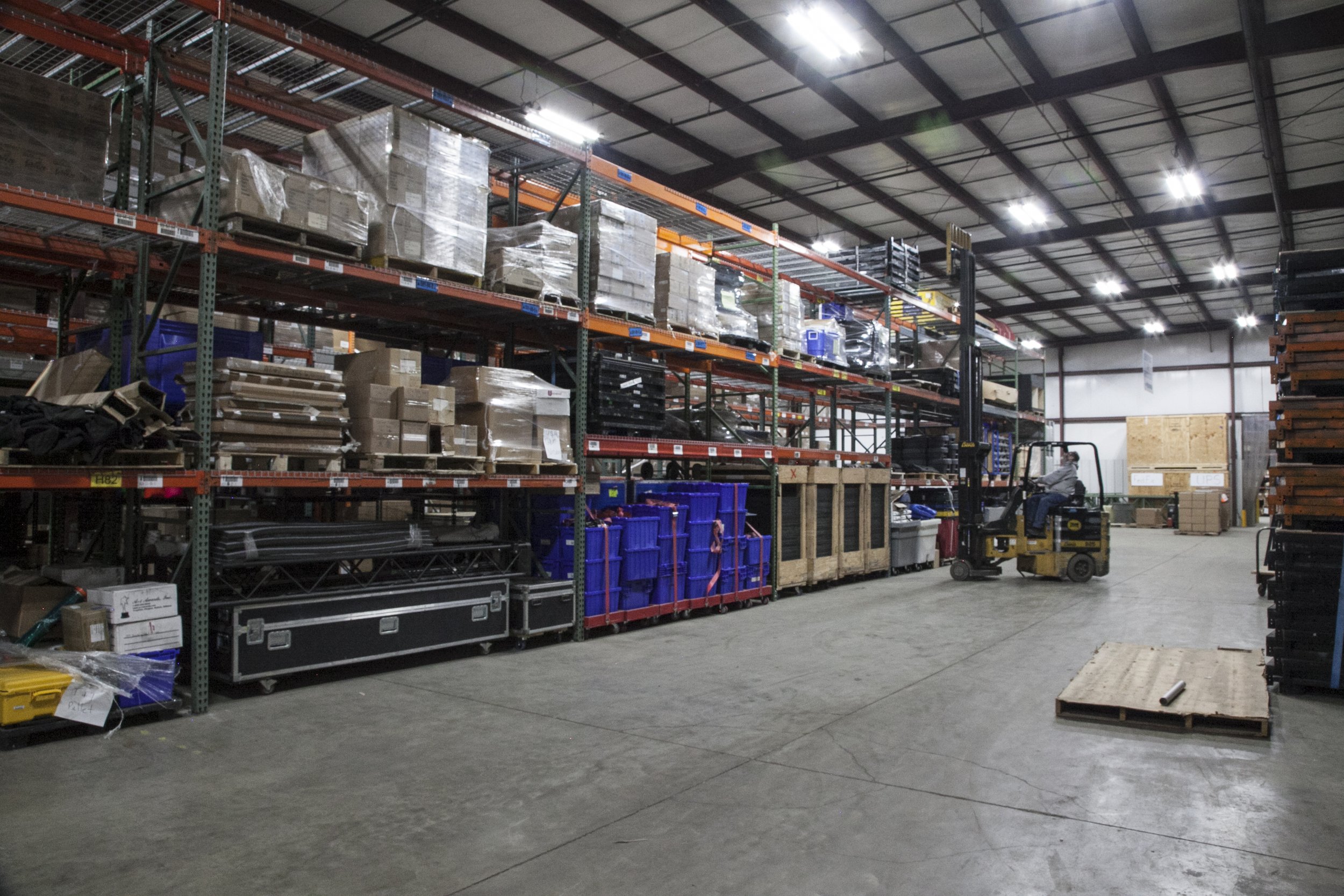 Interior of a warehouse with shelves stacked with various boxes and containers, a person operating a forklift, and an empty wooden pallet on the floor.