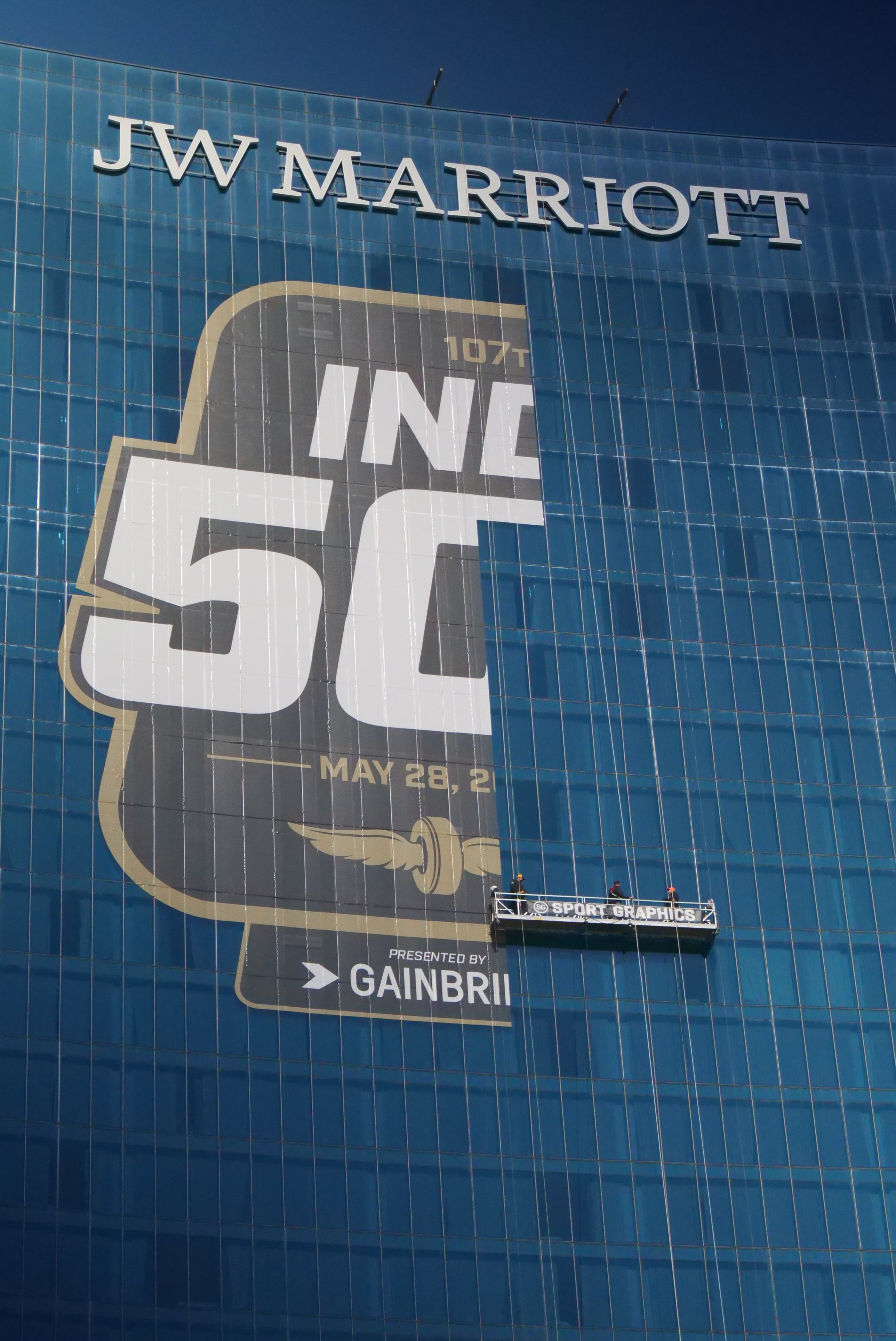 Large advertisement on the JW Marriott building for the Indy 500 race, with workers on a suspended platform.