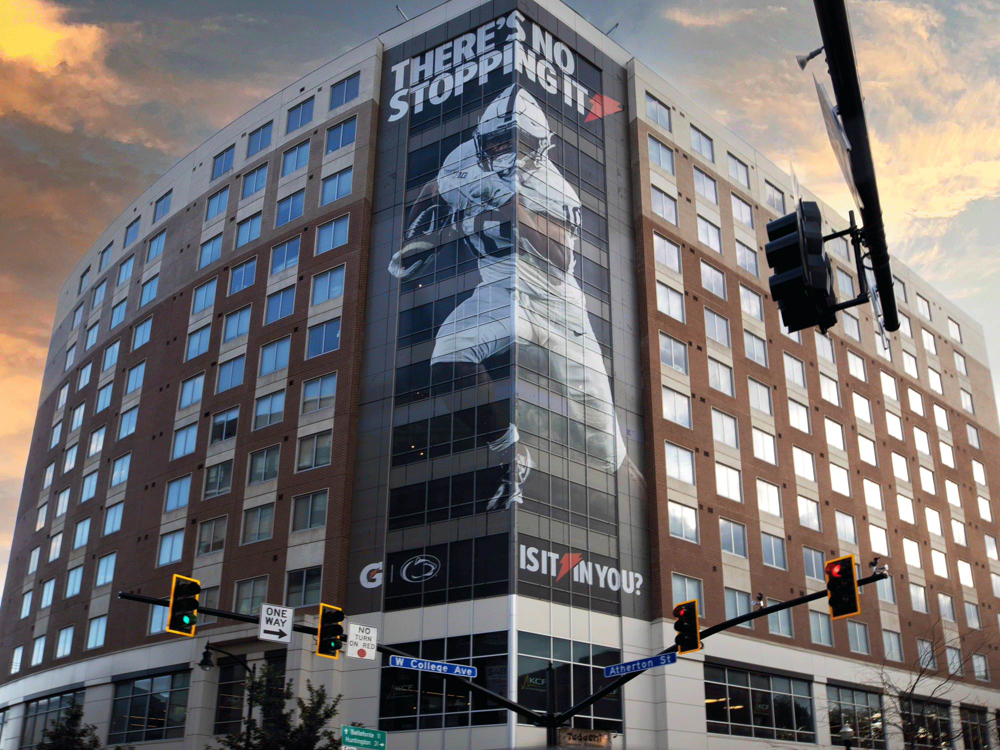 Large building with an advertisement featuring a football player; text reads "There's No Stopping It" and "Is It In You?"; sunset sky in the background; traffic lights and street signs at intersection.