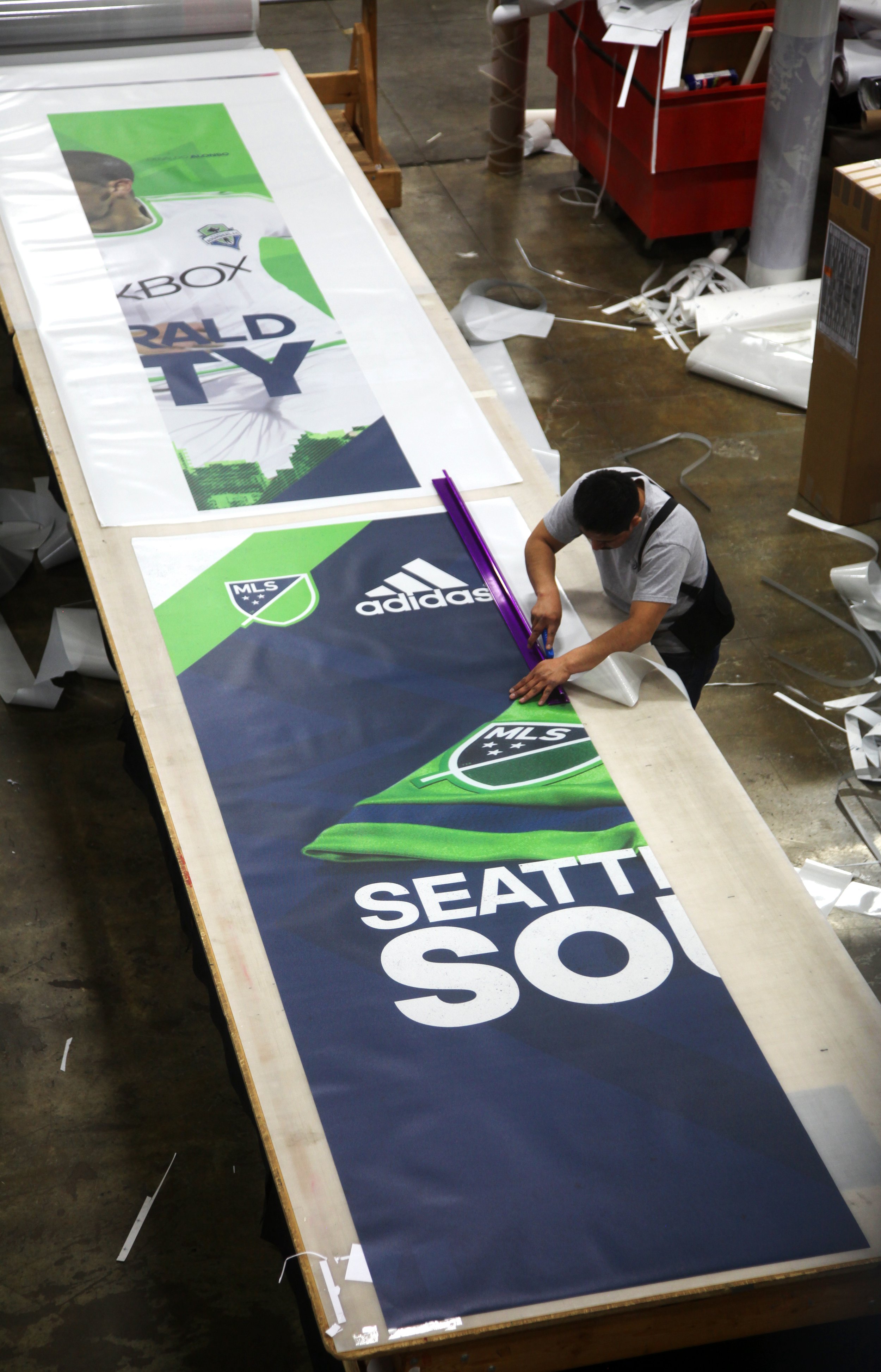 Man preparing large banners for Seattle Sounders with MLS and Adidas logos in a workshop setting.