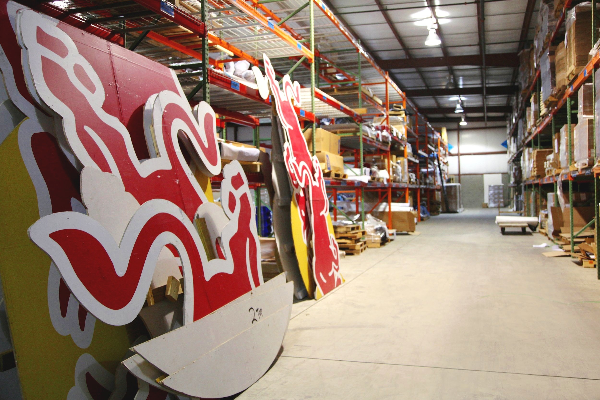 Warehouse interior with large red and white cutout signs, shelves filled with boxes and materials on either side.