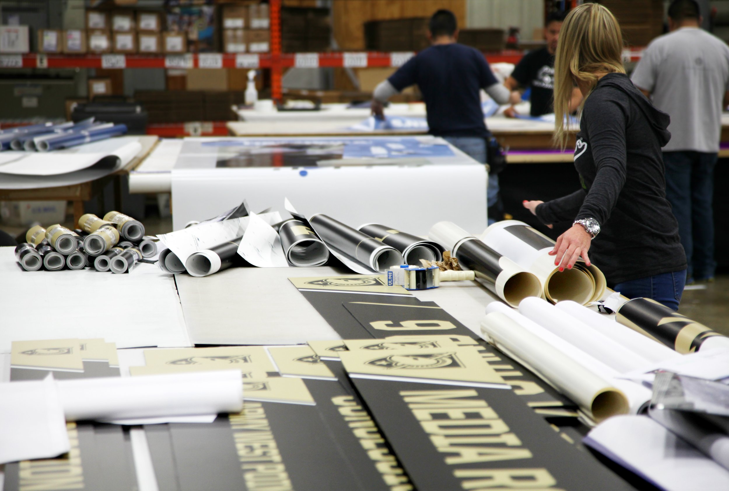 People working in a print shop with rolled and flat poster materials on tables.